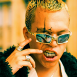 Person with blond hair, wearing reflective sunglasses, rings, and a dark textured jacket, displaying metal grills on teeth and a black face tattoo in an outdoor urban setting.