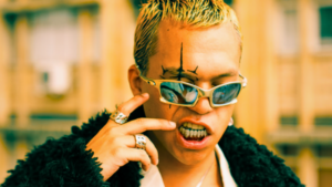 Person with blond hair, wearing reflective sunglasses, rings, and a dark textured jacket, displaying metal grills on teeth and a black face tattoo in an outdoor urban setting.