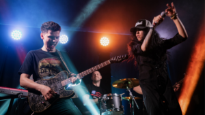 A guitarist, drummer, and singer perform energetically on stage under colorful lights.