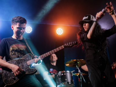 A guitarist, drummer, and singer perform energetically on stage under colorful lights.