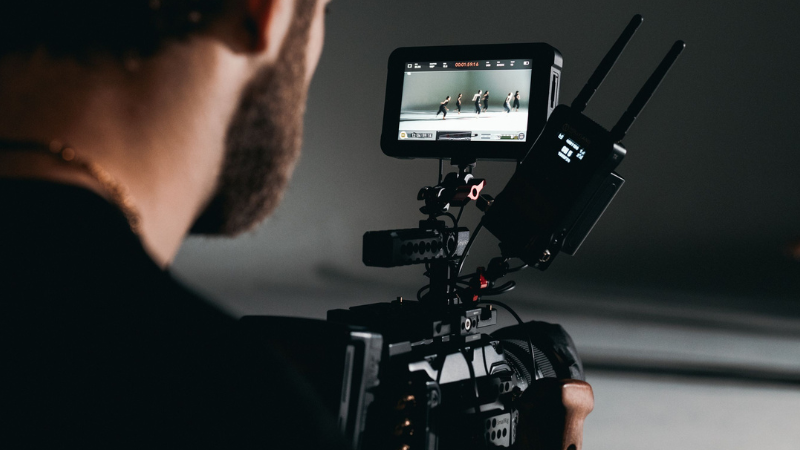 A person operates a professional video camera, focusing on a small monitor displaying dancers in motion during a live video recording.