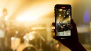 A person records a live band performance on a smartphone; the band and stage are visible through the phone screen in a dimly lit venue, capturing content perfect for YouTube monetization for musicians.