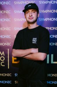 A team member wearing a black T-shirt and cap stands with arms crossed in front of a backdrop with repeated "SYMPHONIC" text.