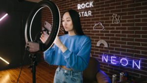 A woman adjusts a smartphone on a ring light in a room with exposed brick walls and neon signs reading "ROCK STAR" and "NEON," preparing to create Top Fan Videos.