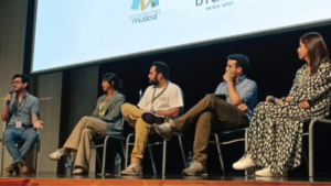 Five people sit on a stage panel, four listening while one speaks into a microphone; behind them, a presentation screen displays information about CAMM 2026.