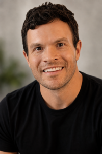 Smiling man with short dark hair and a black t-shirt sits in front of a blurred gray background.
