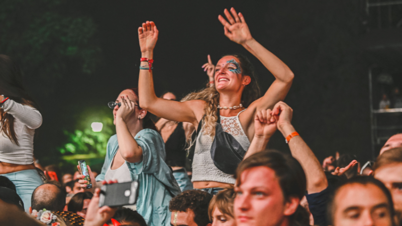 People at a crowded outdoor concert, some sitting on shoulders, enjoying the event; Amazon Music Delivered brings energy as a woman in face paint waves and smiles in the center.