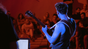 A musician with a guitar performs on stage facing a seated audience under reddish lighting, showcasing his talent and aiming to monetize music in global markets.