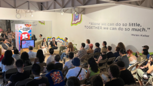 A seated audience listens to a panel discussion in a brightly lit room; a Helen Keller quote is displayed on the wall behind the speakers.