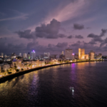 A city skyline at dusk with buildings lit up along a curved waterfront, reflecting lights on the water under a cloudy purple sky—capturing the vibrant energy of All About Music Mumbai.