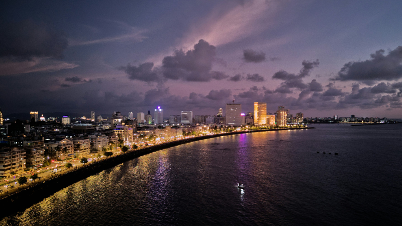 A city skyline at dusk with buildings lit up along a curved waterfront, reflecting lights on the water under a cloudy purple sky—capturing the vibrant energy of All About Music Mumbai.