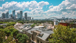 A cityscape featuring modern high-rise buildings, leafy residential areas, and the Sydney Harbour Bridge under a partly cloudy sky, capturing the creative spirit of Indie-Con in a vibrant urban setting.