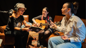 Three women sit together indoors; one holds a notepad, one plays an acoustic guitar, and one speaks or sings while holding a phone. Audio equipment is visible.