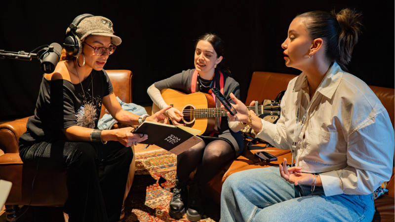 Three women sit together indoors; one holds a notepad, one plays an acoustic guitar, and one speaks or sings while holding a phone. Audio equipment is visible.