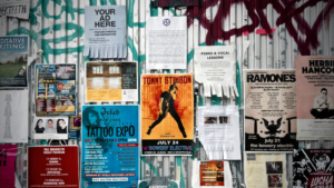 A wall covered with various posters and flyers, including event ads, paid ads for music releases, tattoo expo promotions, and lesson announcements, set against a graffiti-covered metal surface.