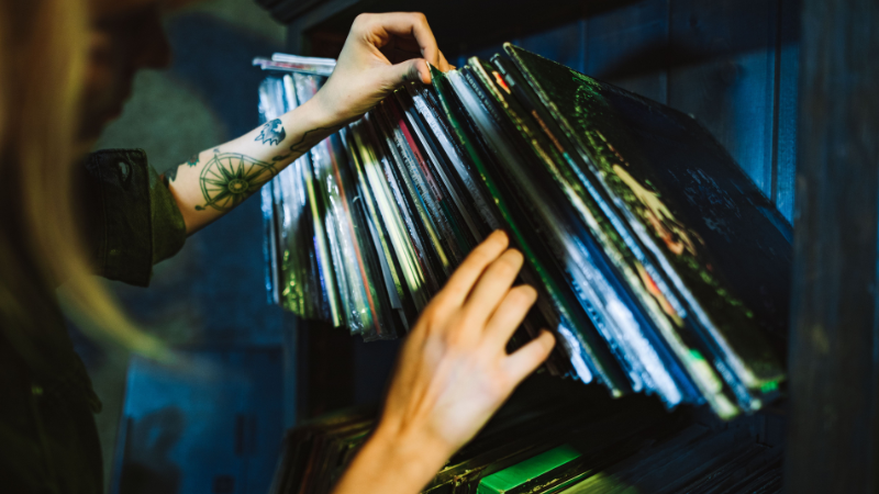 A person with a tattoo on their forearm flips through a collection of vinyl records on a shelf.