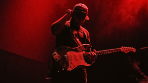 A person wearing a cap plays an electric guitar on stage under red lighting, with their face partly in shadow, perhaps pondering whether platforms like SoundCloud are worth it for independent artists seeking exposure.